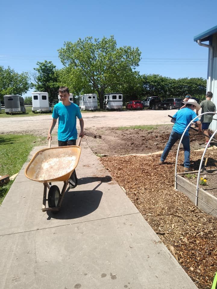 Two people work in a garden, one pushing a wheelbarrow along a sidewalk, with mulch and trees nearby.