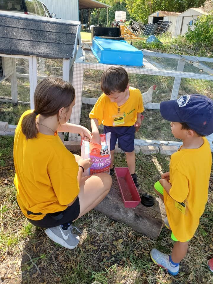 Three children in yellow shirts gather around a small sandbox outdoors, digging with a pink shovel.