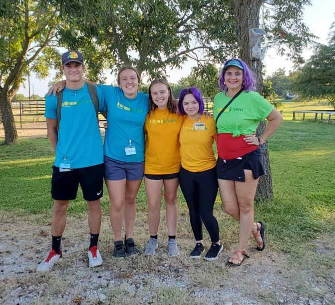 Five people in colorful shirts standing together on a grassy field with trees in the background.