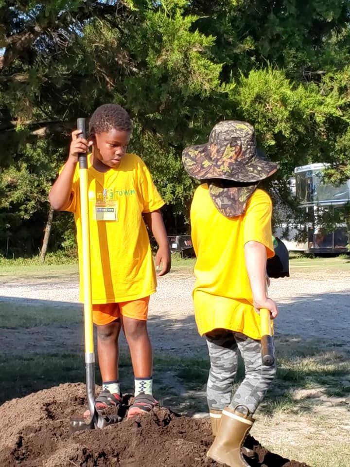 Two children in yellow shirts stand outdoors with shovels near a dirt pile.