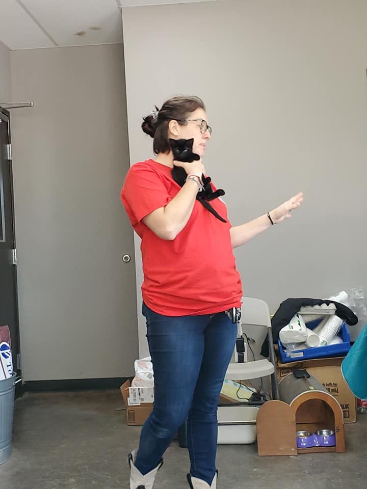 Person in a red shirt speaking into a handheld microphone in a room with boxes and supplies.