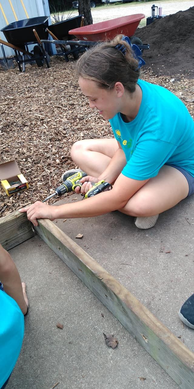 Person kneels on pavement, working with a small object beside a wooden frame and gravel area.