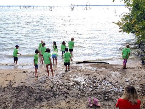 People in green shirts standing on a muddy lakeshore near the water, with trees in the background