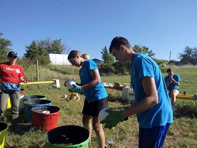Volunteers in blue shirts wash buckets outdoors beside large tubs on a sunny day