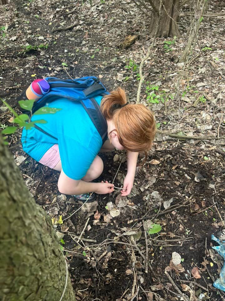 Person kneeling in a wooded area, picking up small items from the ground while wearing a blue shirt and backpack.