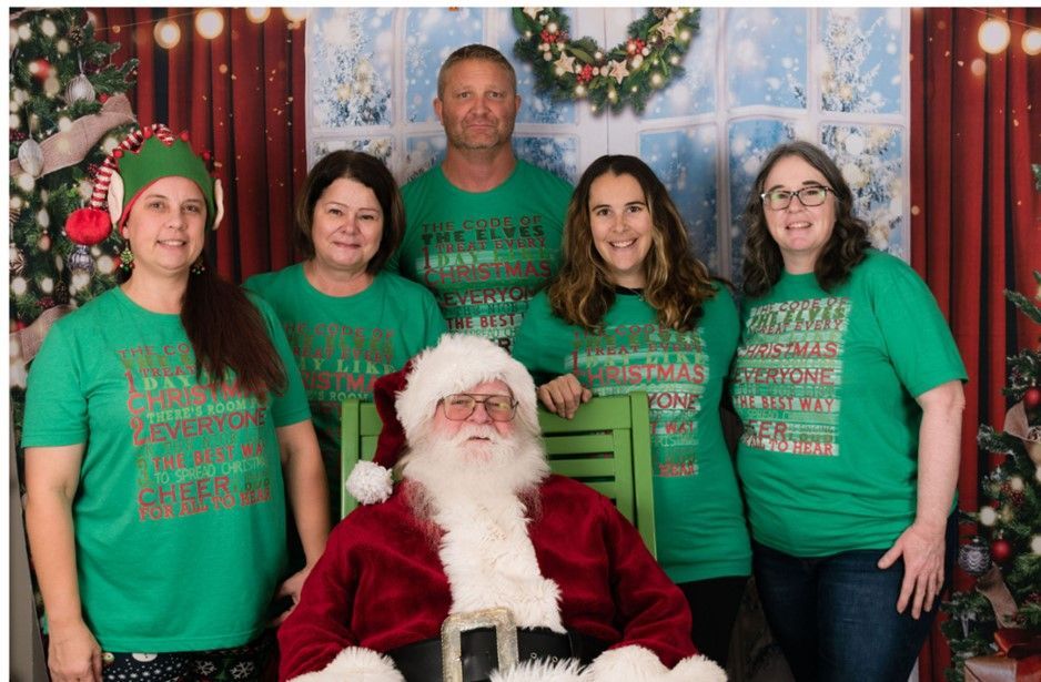 Santa seated with five people in green shirts posing in front of a festive holiday backdrop