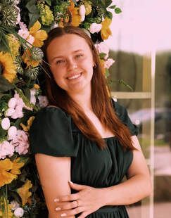 Smiling woman in a dark green dress standing beside a flower-covered arch