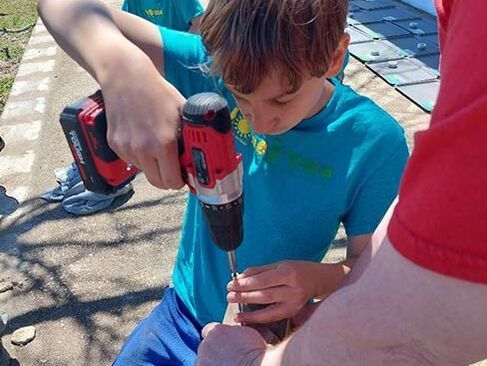 Child using a red cordless drill outdoors while an adult steadies the workpiece.