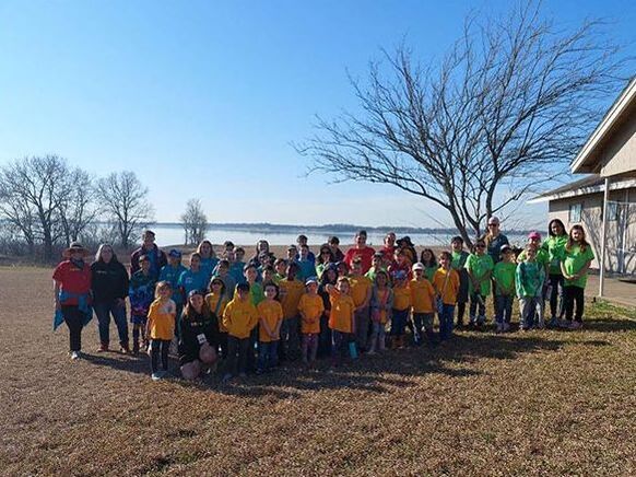 Group of children and adults in colorful coats standing outdoors near a lake on a sunny day