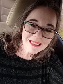 Smiling person wearing glasses and a dark striped top, seated in a car.