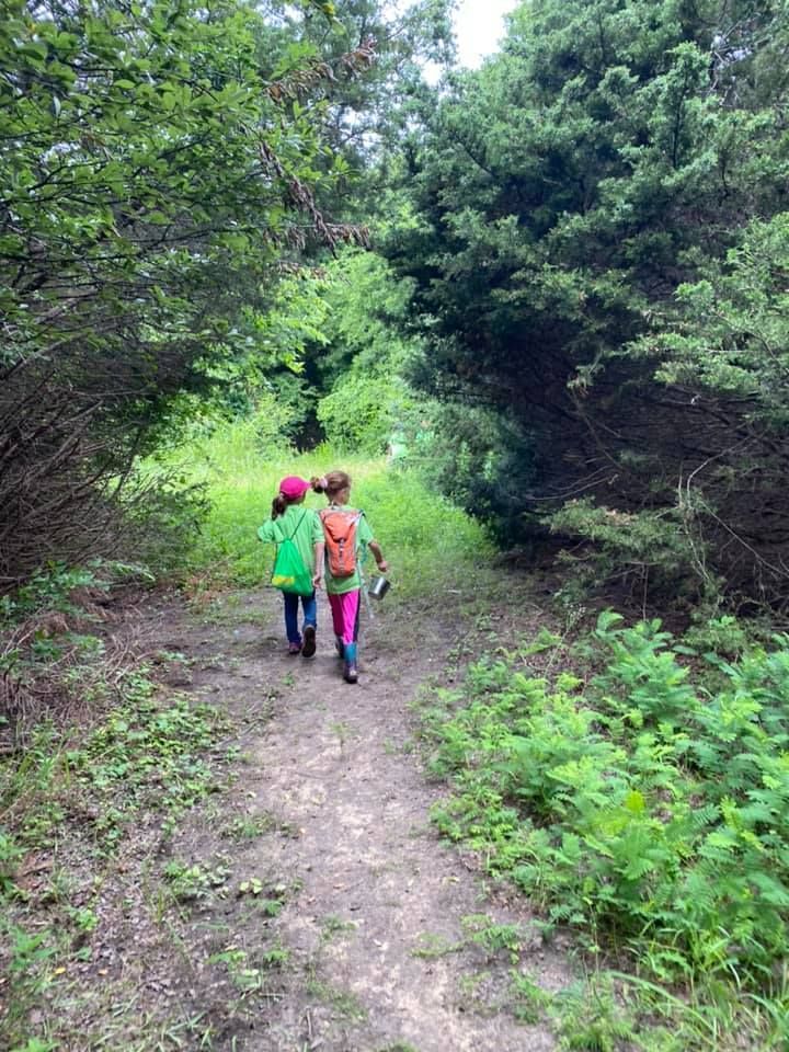 Two children in bright clothes walk along a narrow forest path under green trees.