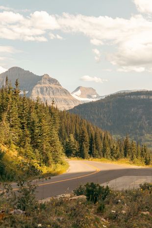 Winding mountain road through a forest with distant peaks under a partly cloudy sky.