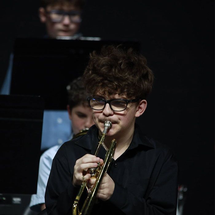 Young musician playing trumpet on stage, with a blurred pianist in the background.