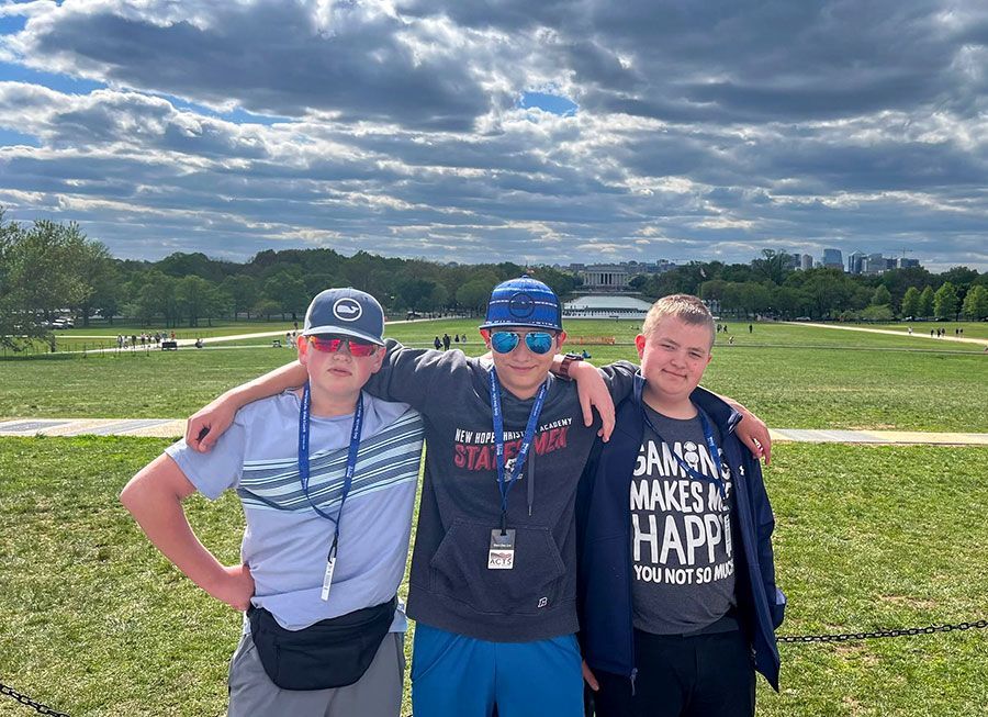 Three friends standing on a grassy field under a cloudy sky, posing with arms around each other.
