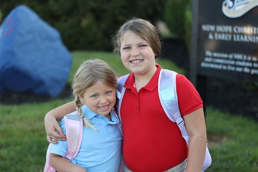 Two children smiling outdoors, wearing red and blue shirts with backpacks.
