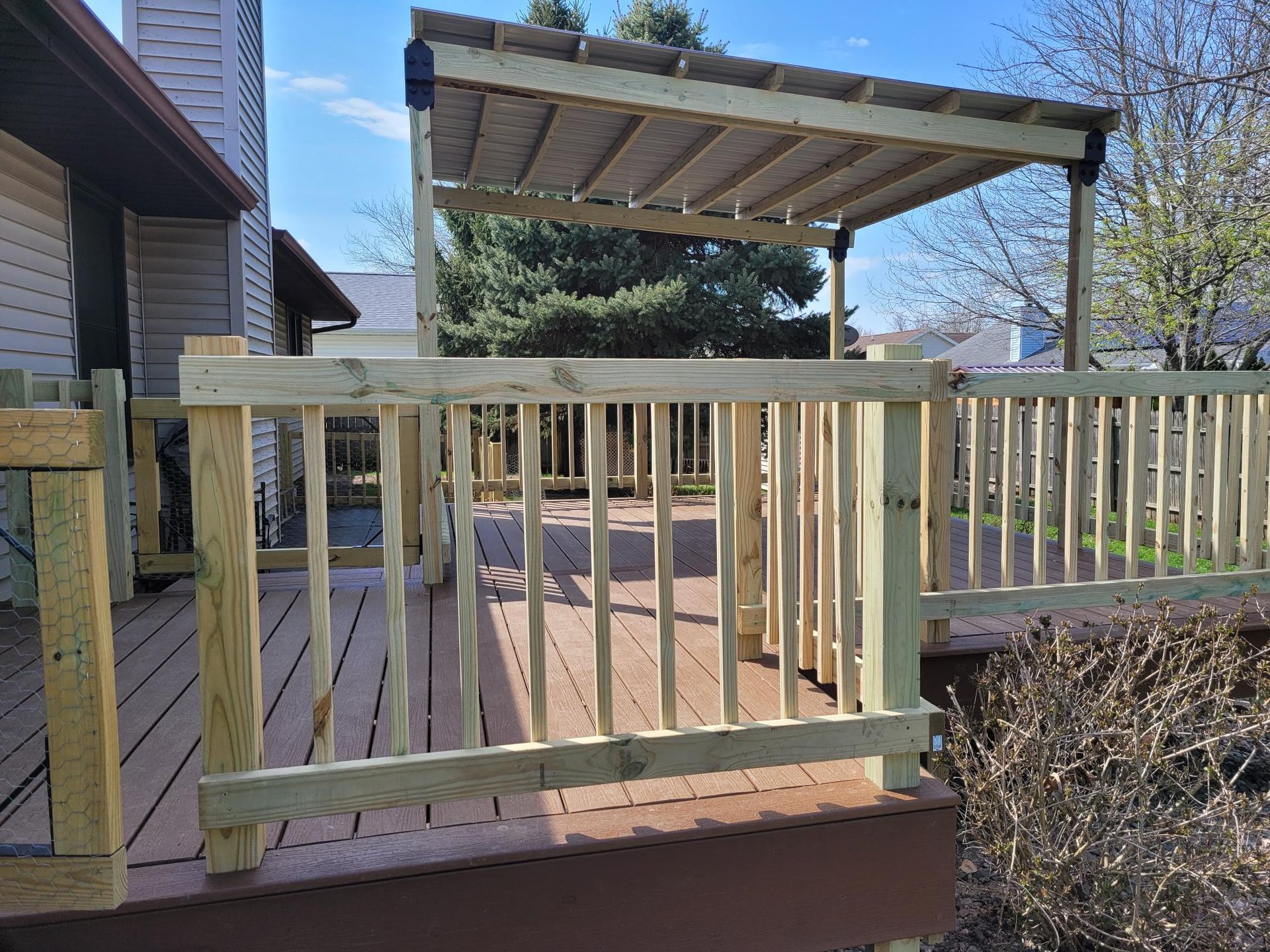 Wooden deck with railings and pergola beside a house, overlooking a yard with leafless shrubs.