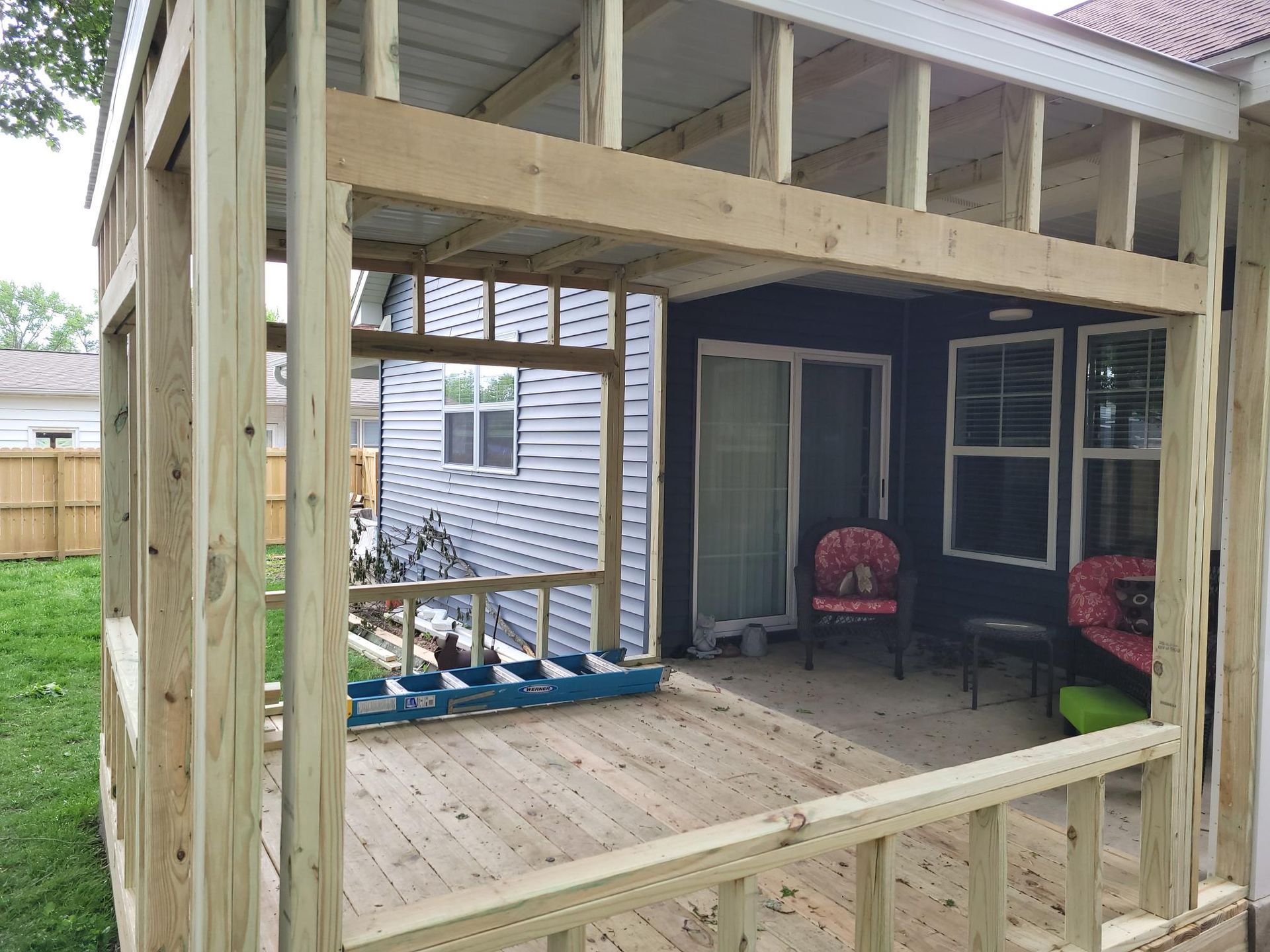 Wooden porch under construction with exposed framing and two red chairs on a covered patio.
