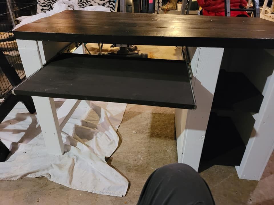 Black desk with a pull-out tray and white legs in a cluttered room with paper on the floor