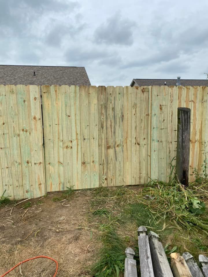 Wooden fence under construction with dirt ground, cloudy sky, and lumber stacked in foreground