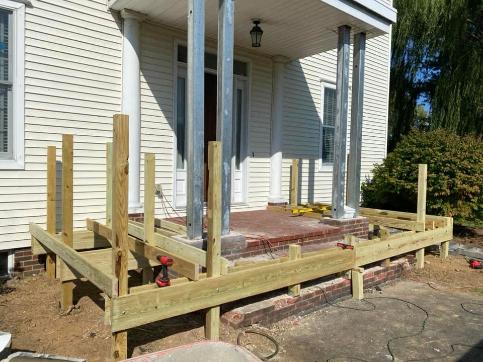 Raised wooden porch frame under construction beside a white house, with posts and beams installed