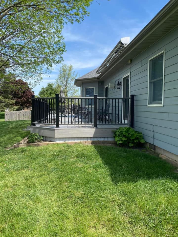 Backyard deck with black railing beside a light gray house and green lawn under a sunny sky