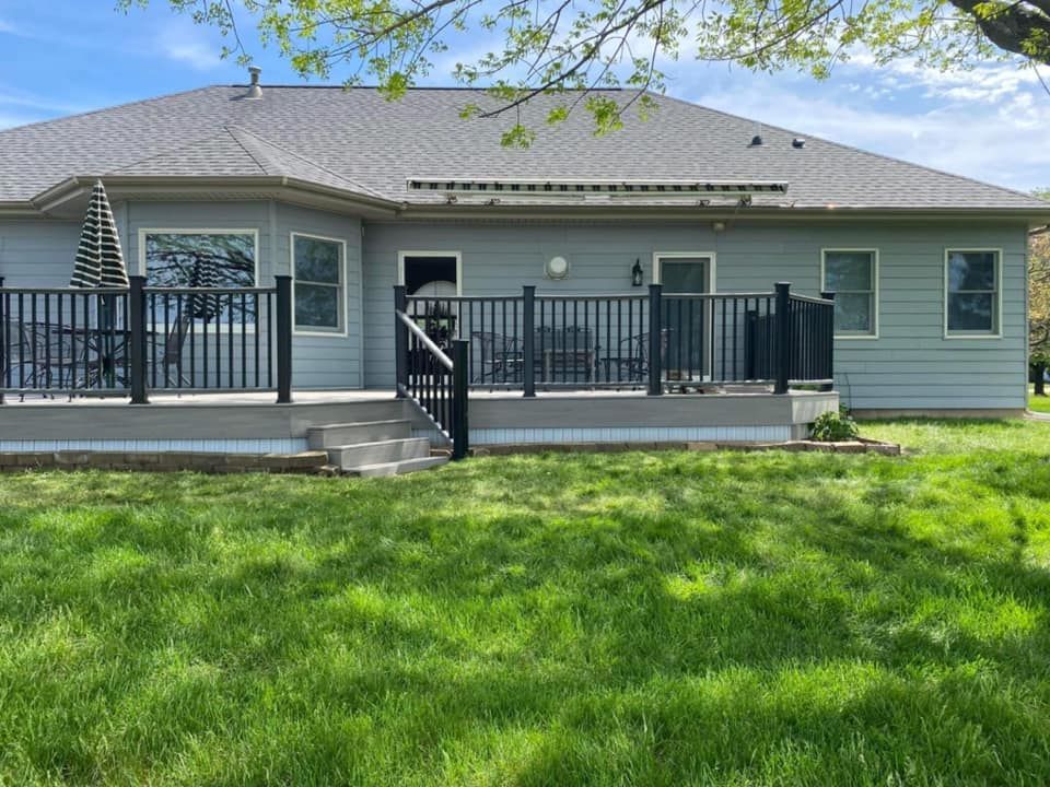 Single-story gray house with front porch, black railing, and green lawn under a bright sky