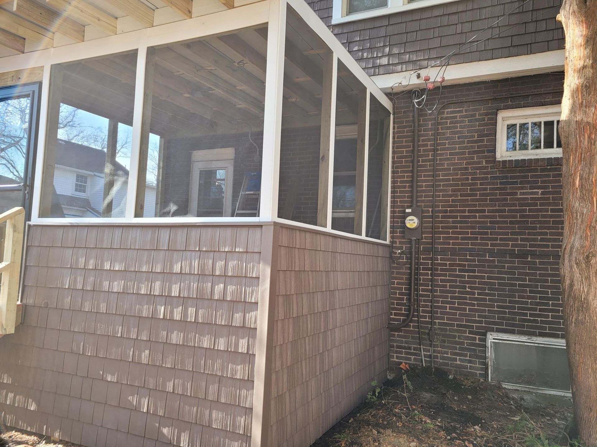 Covered porch with white posts and brown siding beside a brick house, viewed from the side.