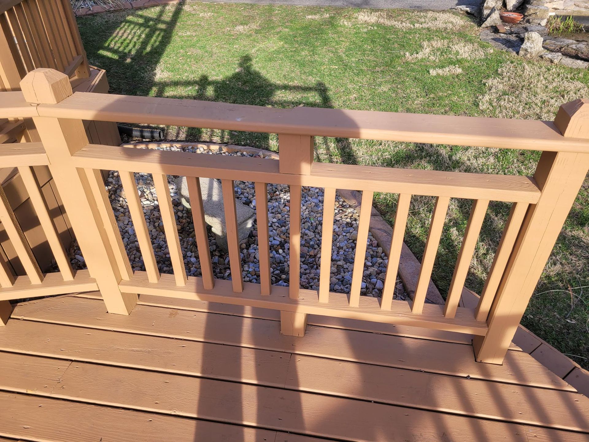 Wooden deck railing with tan posts and brown floorboards overlooking a yard and garden bed.