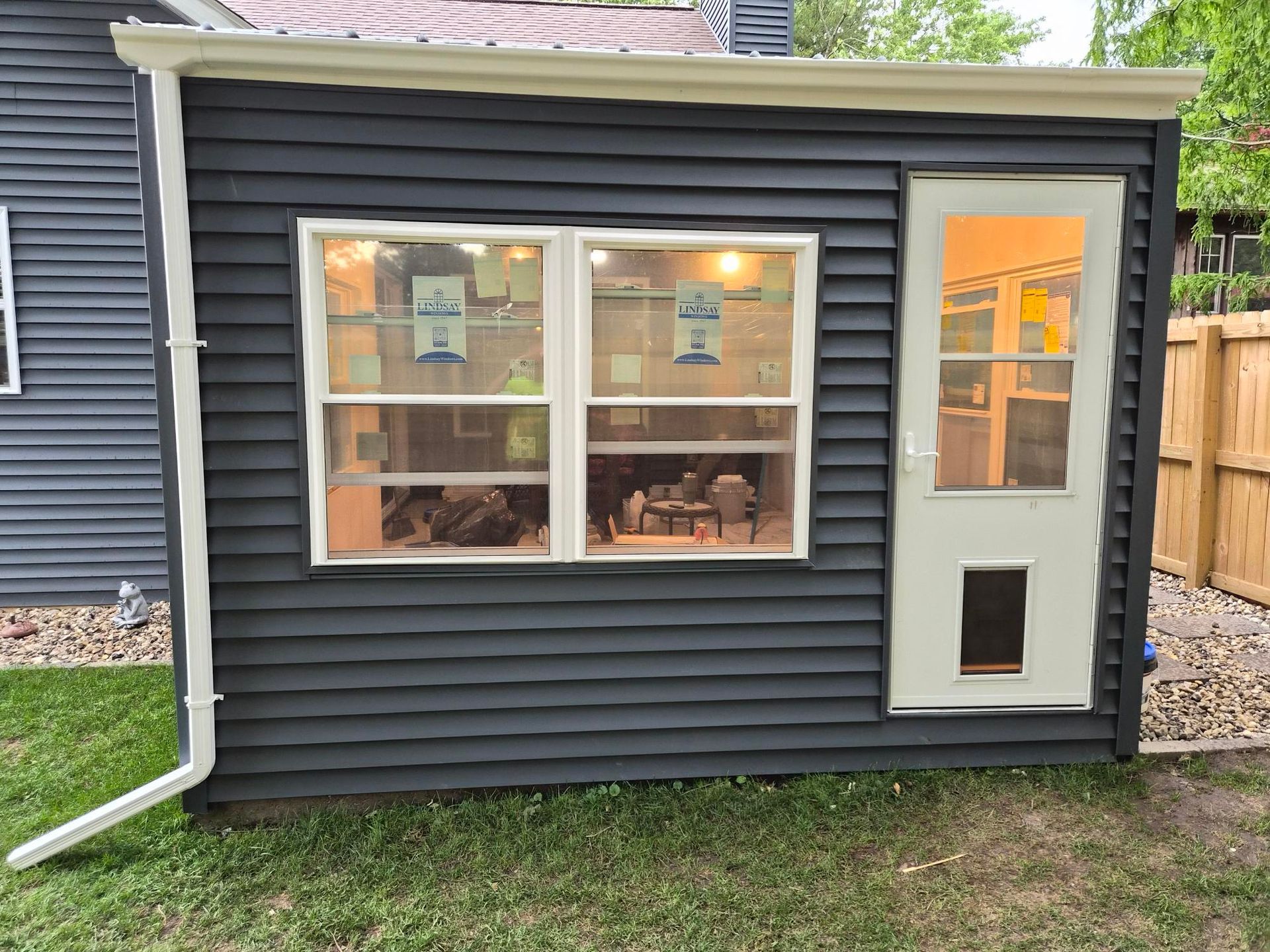 Small dark-gray backyard shed with white-trim windows and door, lit interior, beside a wooden fence and grass.