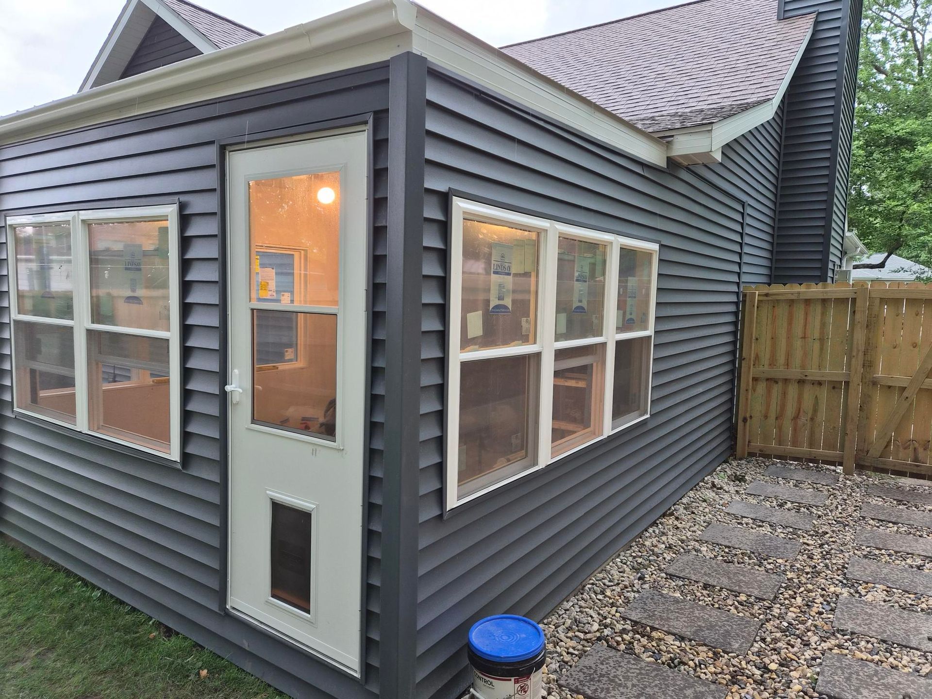 Small gray house addition with white-trim windows, a door, and a paved courtyard beside a wooden fence.