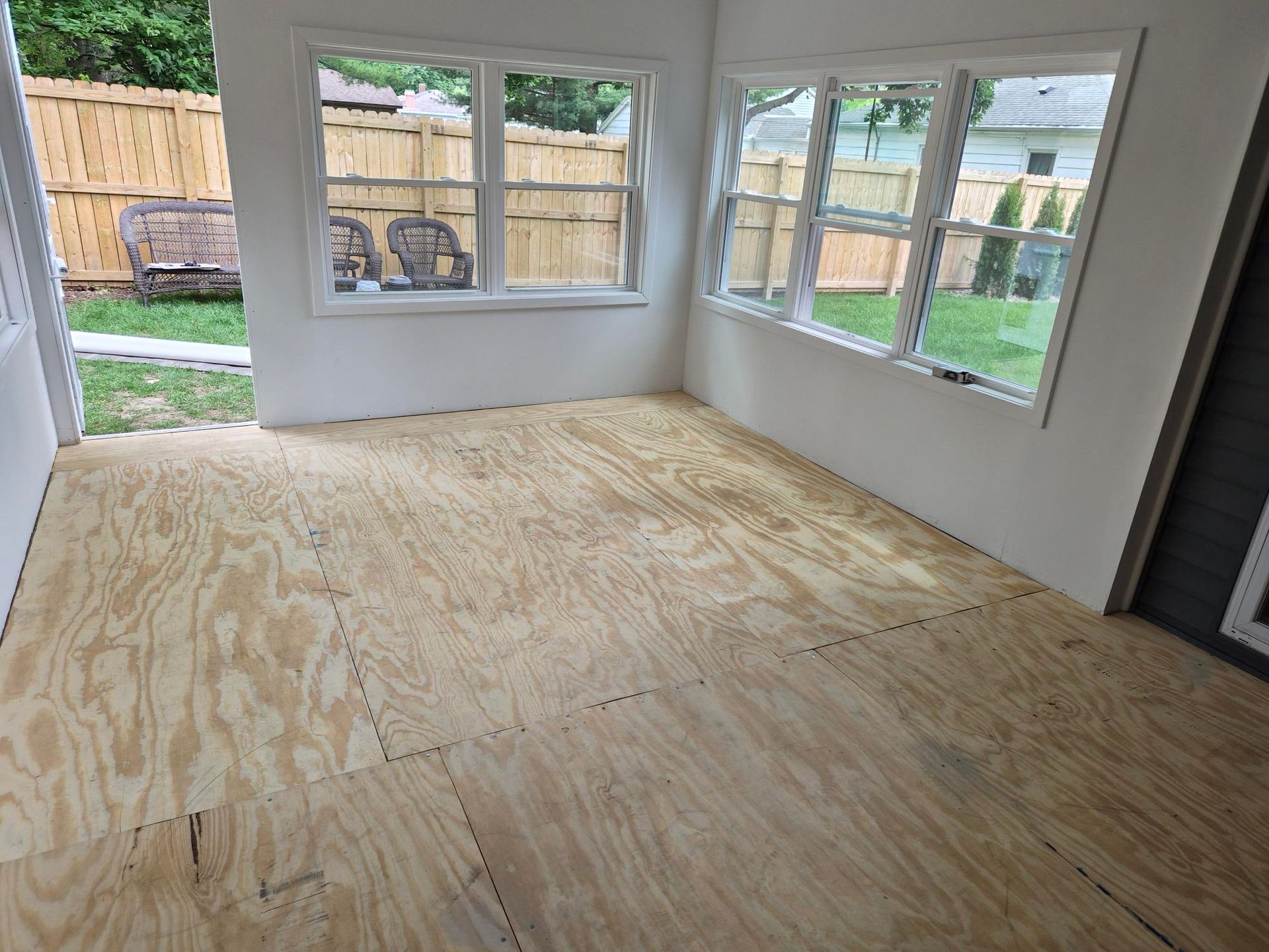 Empty room with unfinished plywood floor and large windows overlooking a fenced yard