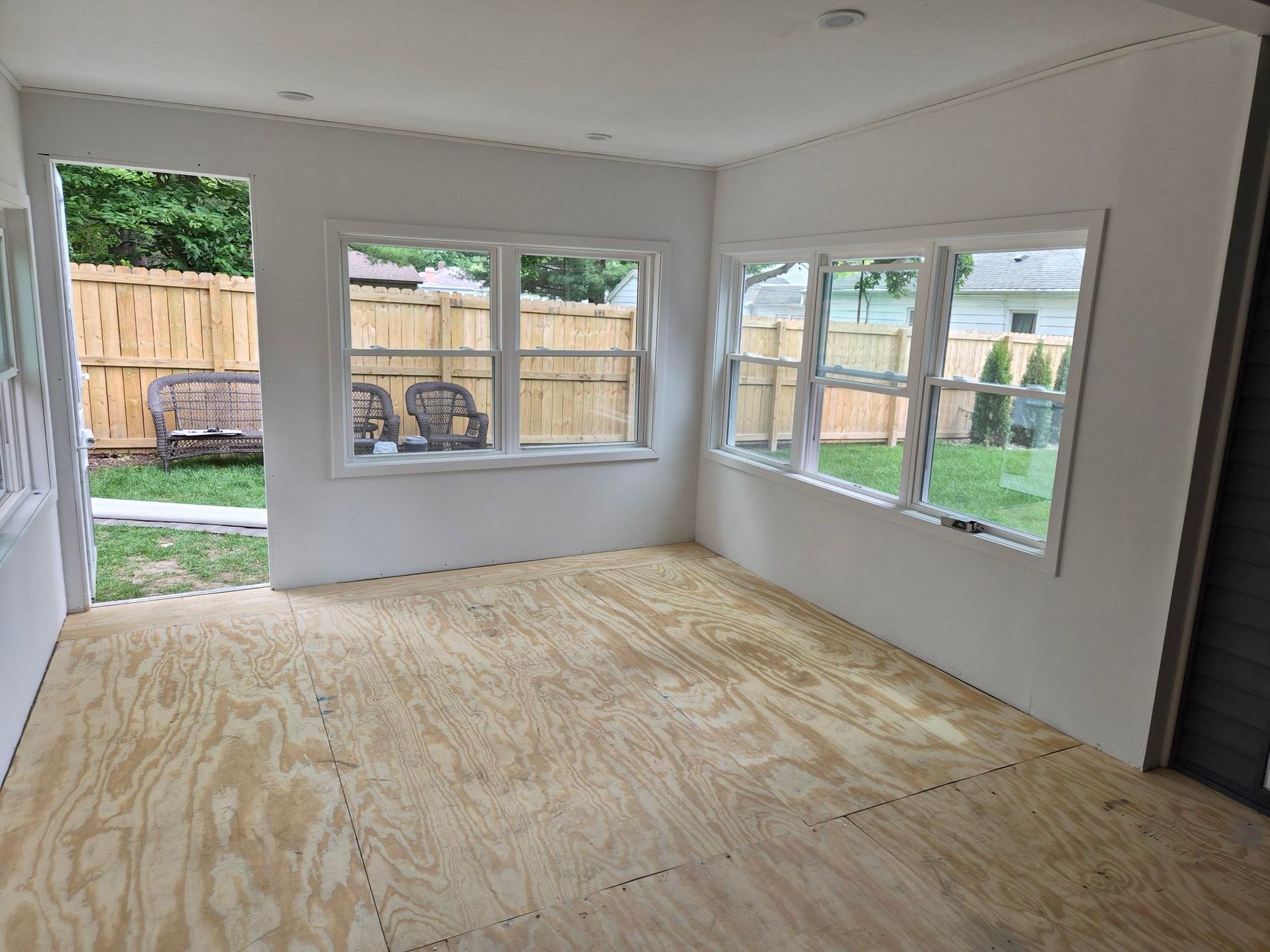 Empty room with plywood subfloor, white walls, and large windows overlooking a fenced backyard.