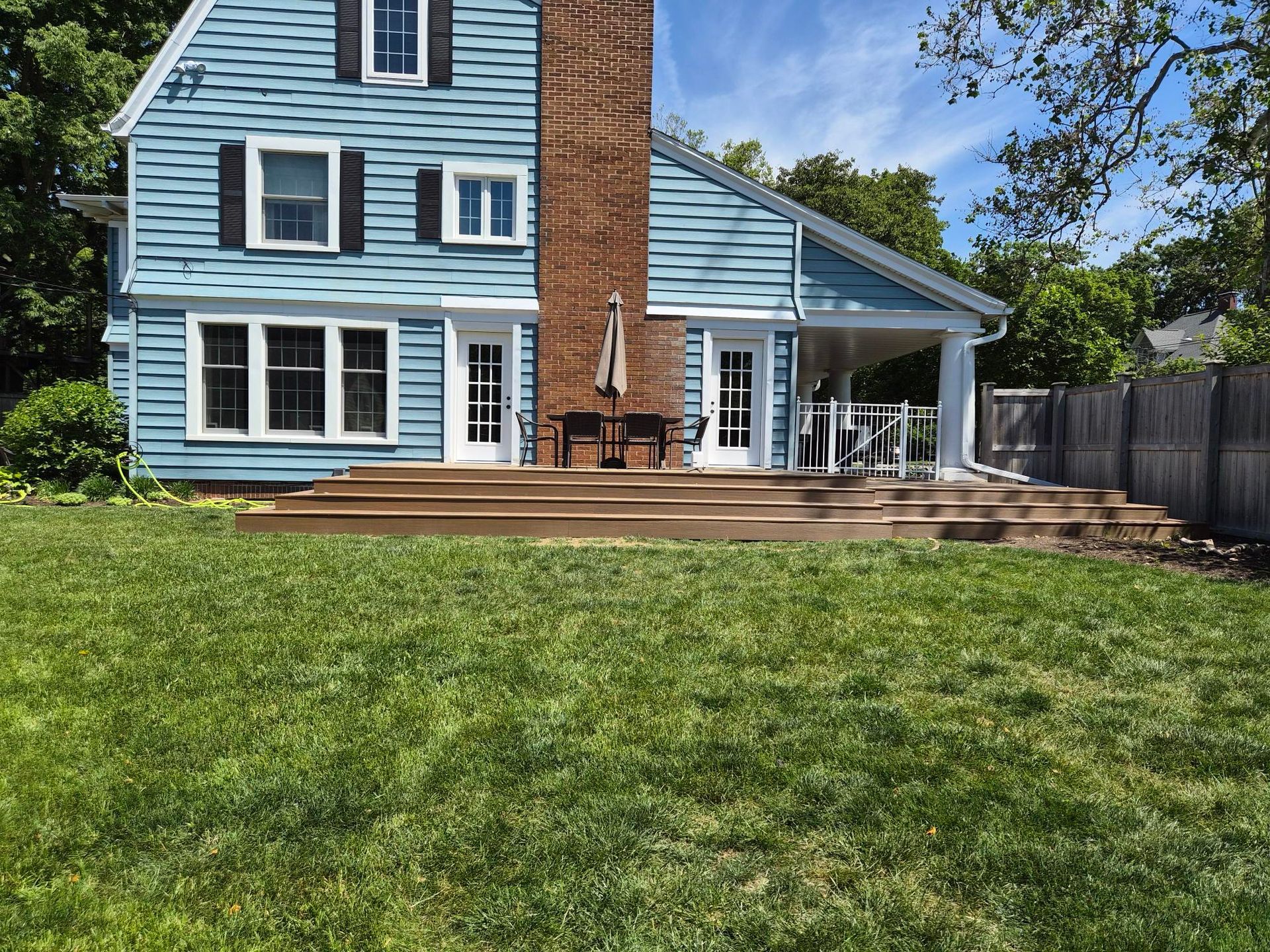 Blue house with white trim, brick chimney, and wooden deck beside a fenced backyard lawn.