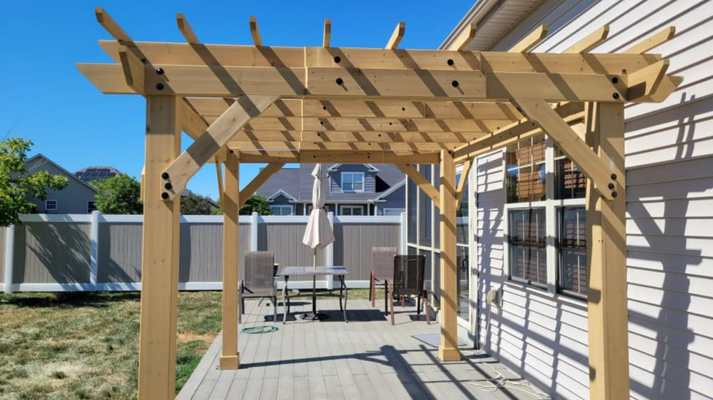 Wooden pergola over a backyard patio with table, chairs, and string lights beside a house