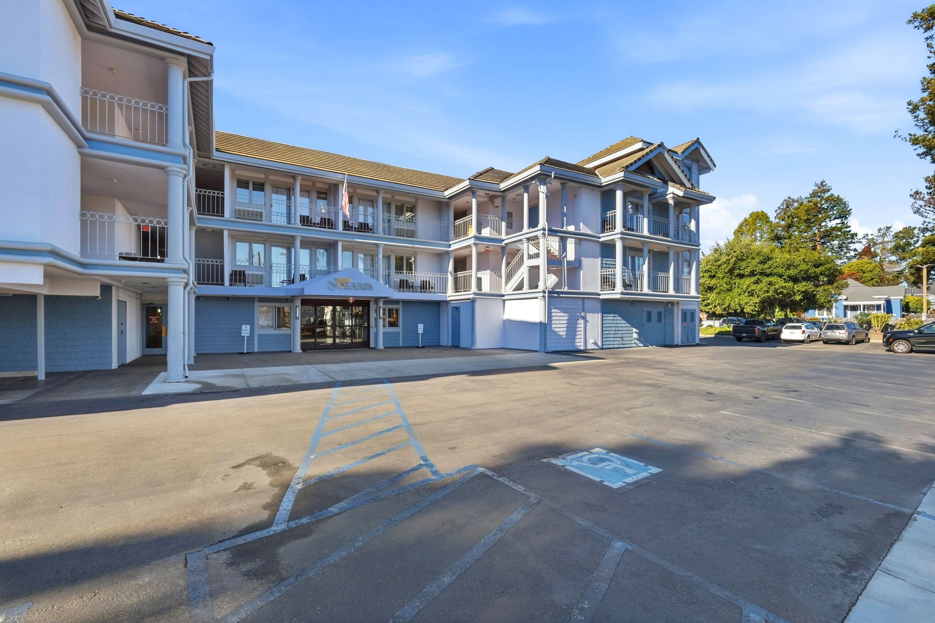 A three-story blue hotel building with white trim, balconies, and a paved parking lot under a clear blue sky.