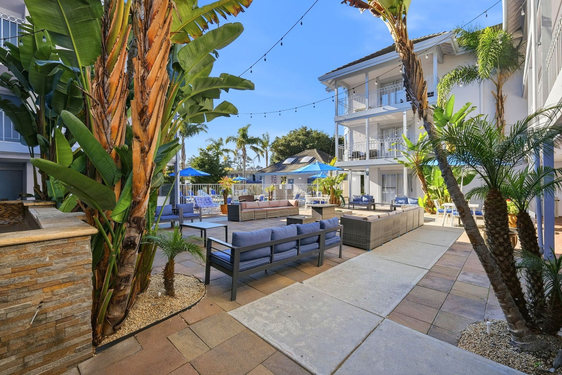 A sunlit hotel courtyard featuring outdoor lounge seating, palm trees, blue umbrellas, and a multi-story white building.