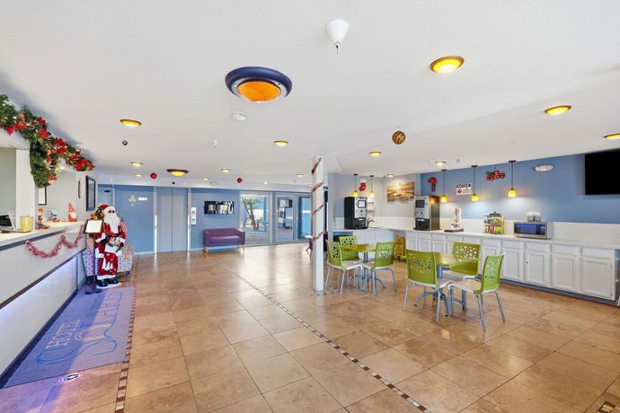 A bright hotel lobby with beige tile floors, a reception desk with Santa decor, and green chairs near a refreshment area.