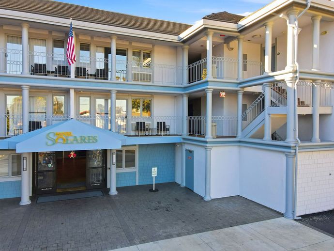 A two-story hotel building with a blue awning, white pillars, balconies, and an American flag flying out front.