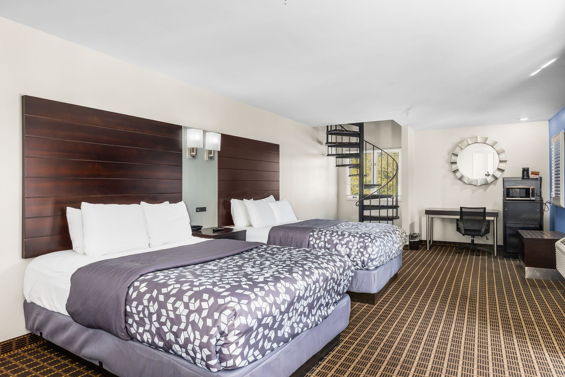 A hotel room with two queen beds, dark wood headboards, patterned carpet, a desk, and a spiral staircase.