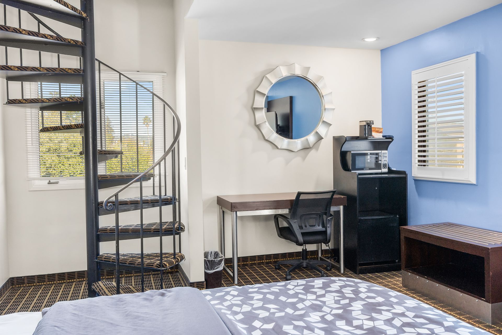 A bedroom with a spiral staircase, a desk with an office chair, a mirror, and a black storage unit against a blue wall.