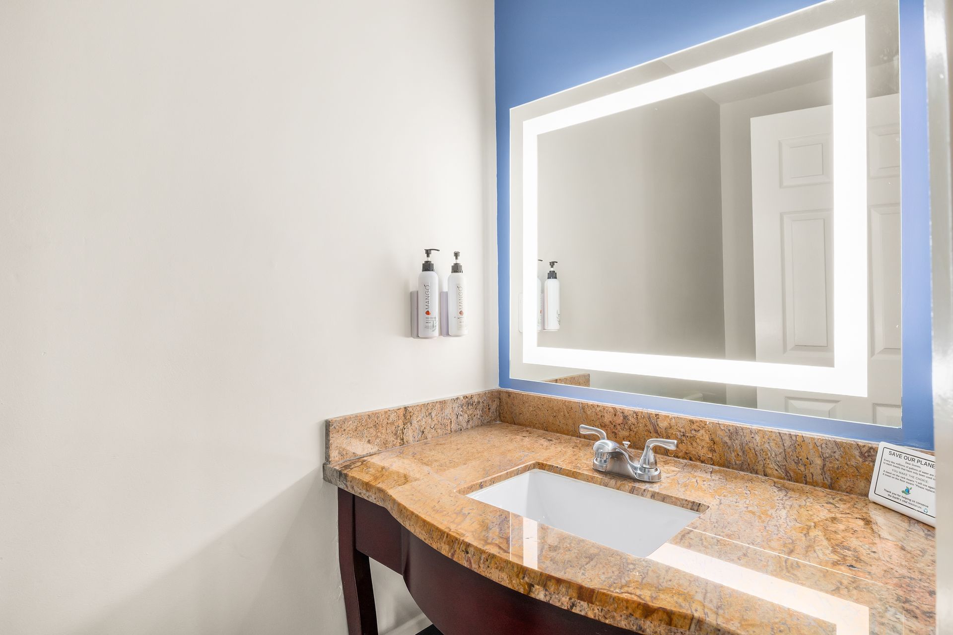 A hotel bathroom vanity with granite countertop, sink, and an illuminated mirror on a blue accent wall.