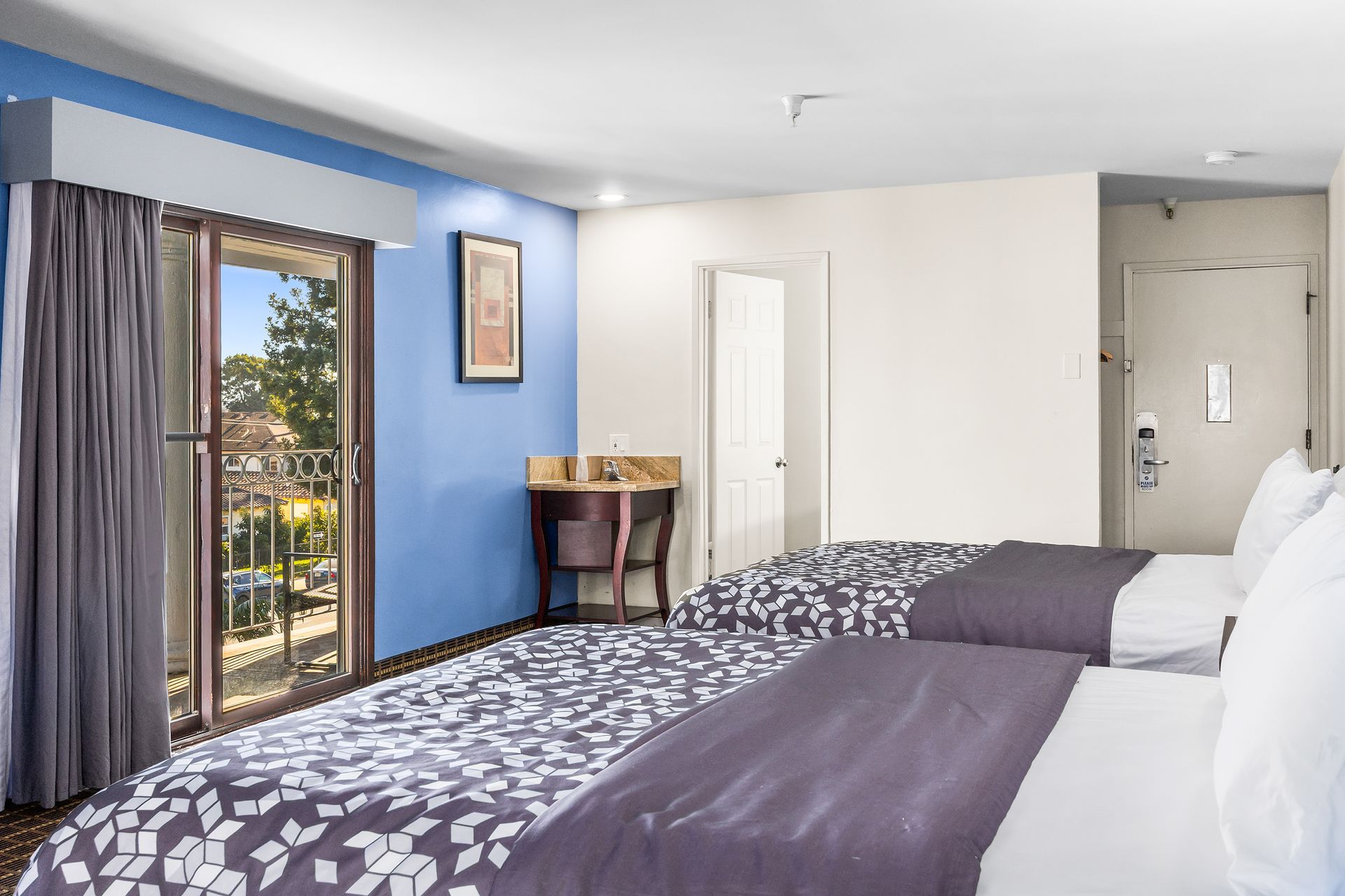 A hotel room with two beds, a blue accent wall, a glass balcony door, and a small desk against the wall.