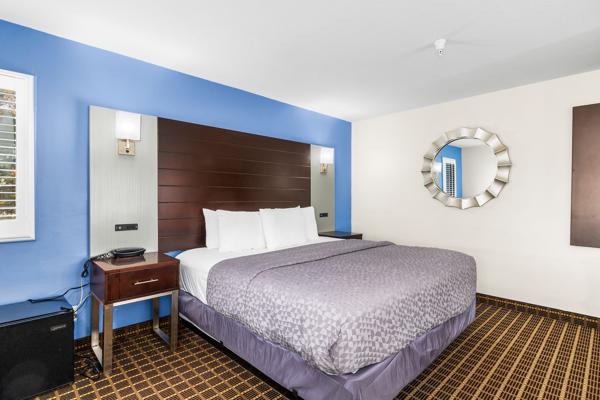 A hotel room featuring a king bed with gray bedding, a dark wood headboard, blue accent wall, and a round wall mirror.