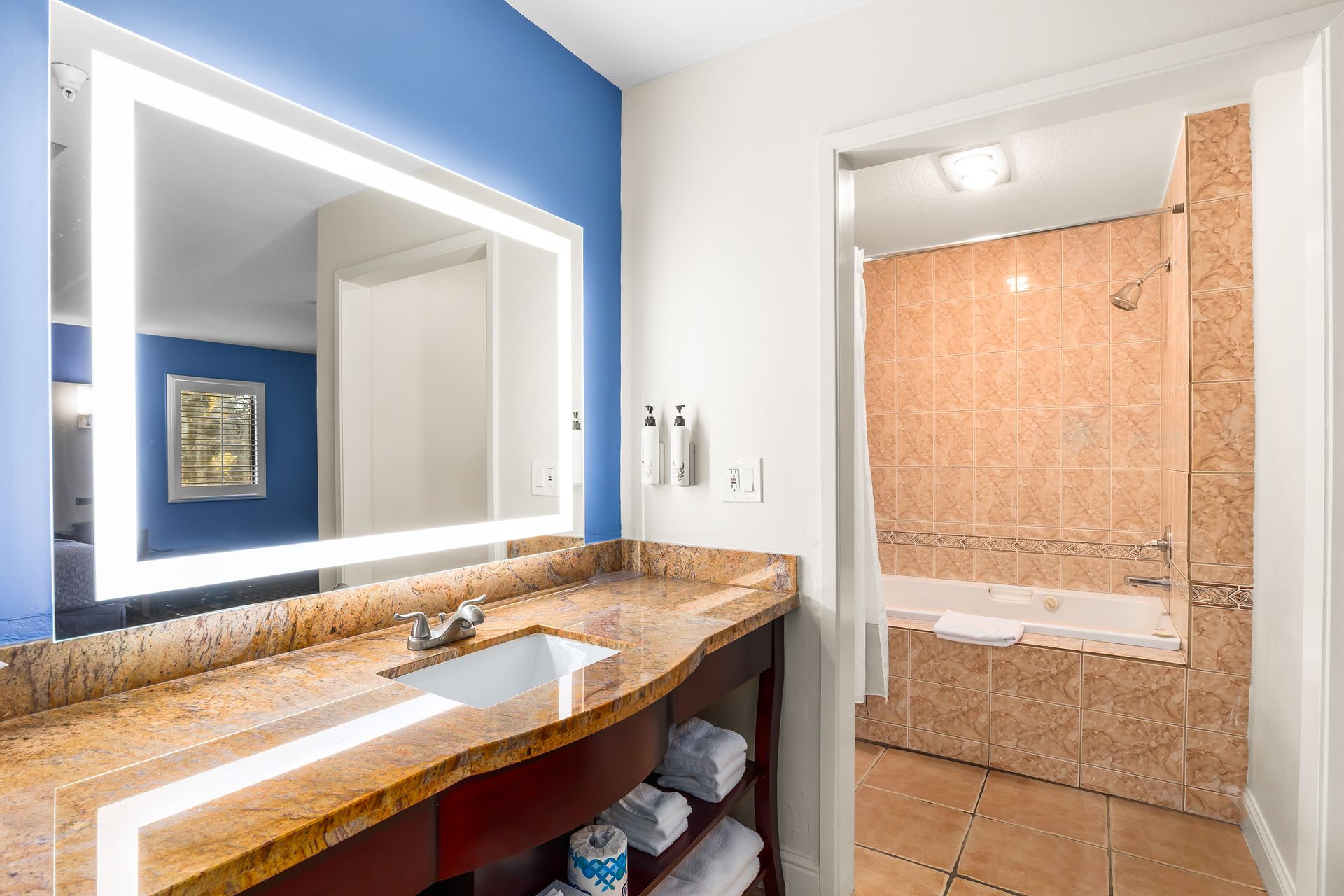 A modern bathroom with a light-up mirror above a tan granite vanity, featuring an adjoining walk-in tiled shower.