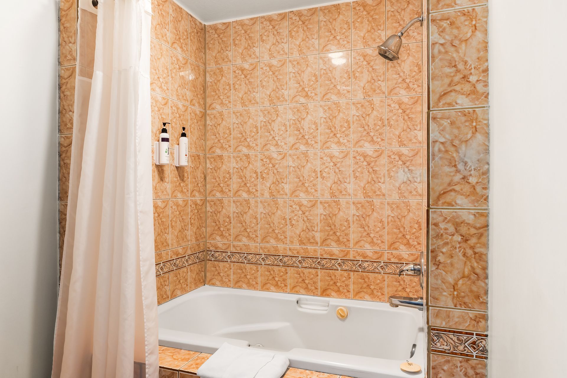 A bathtub with a shower head and light orange, patterned tile walls, and a white shower curtain hanging to the left.