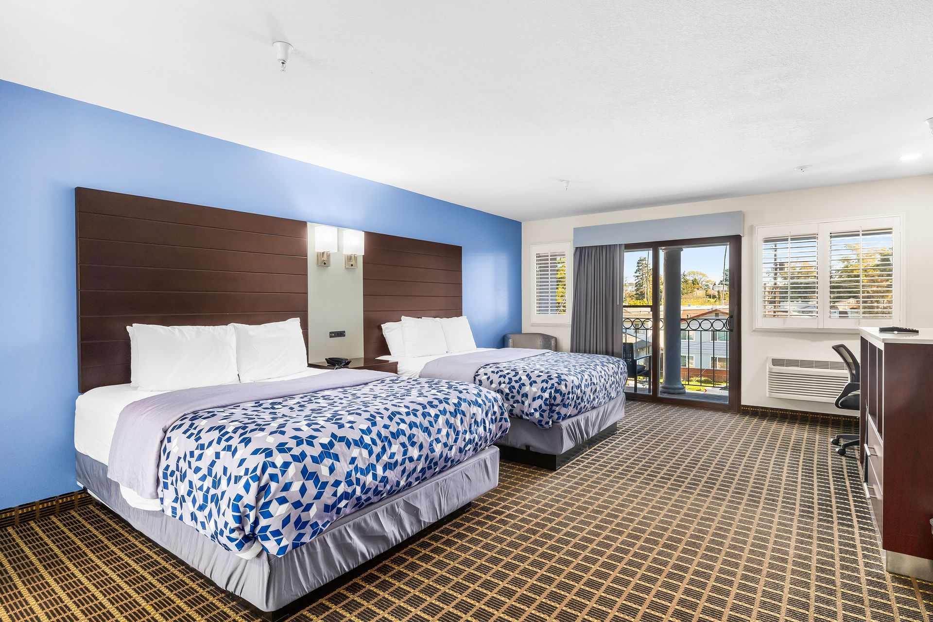 A hotel room with two queen beds featuring blue patterned quilts, a dark wood headboard, and a patio door.