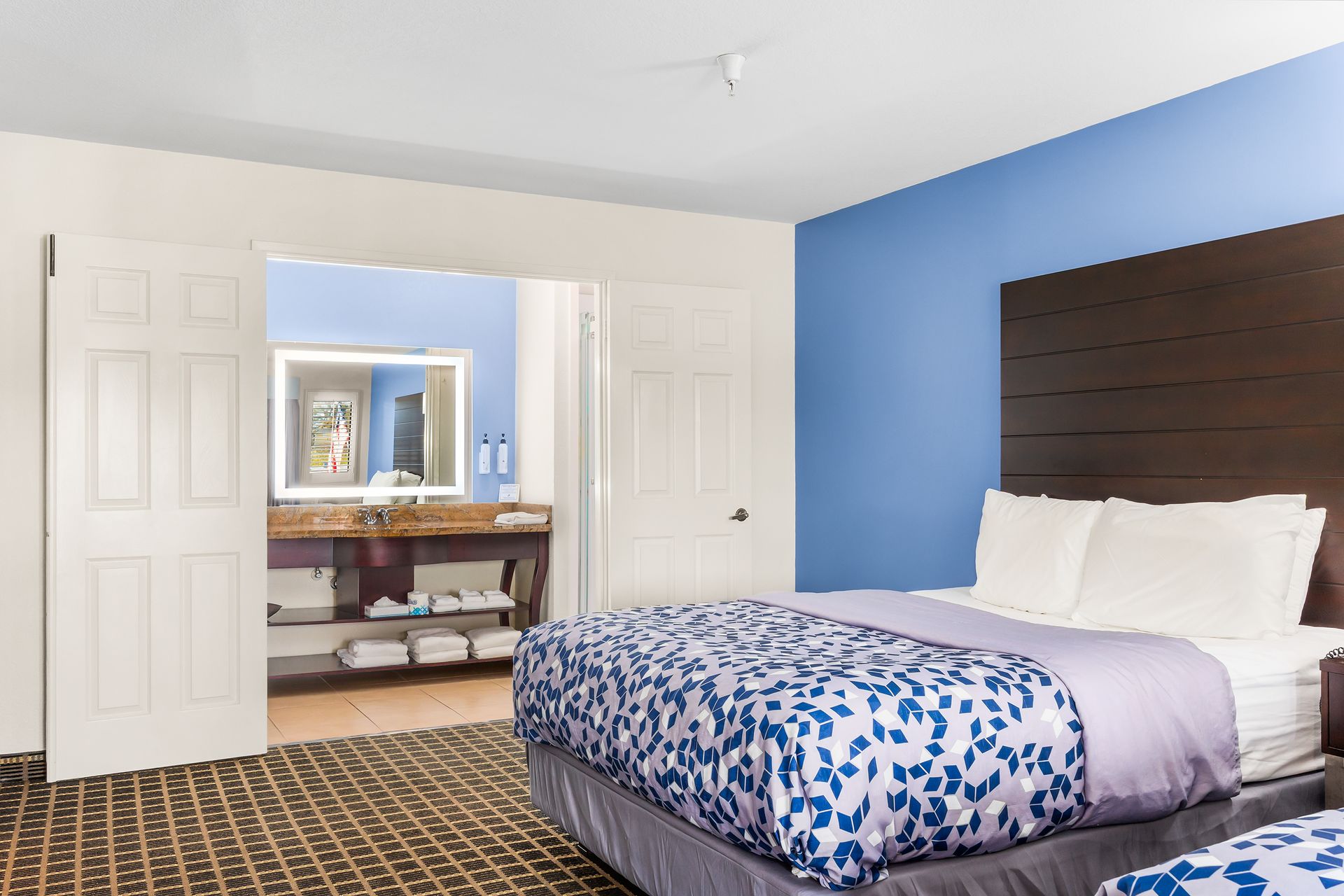 A hotel room with a made bed, patterned comforter, dark wood headboard, blue accent wall, and an open door to a bathroom.