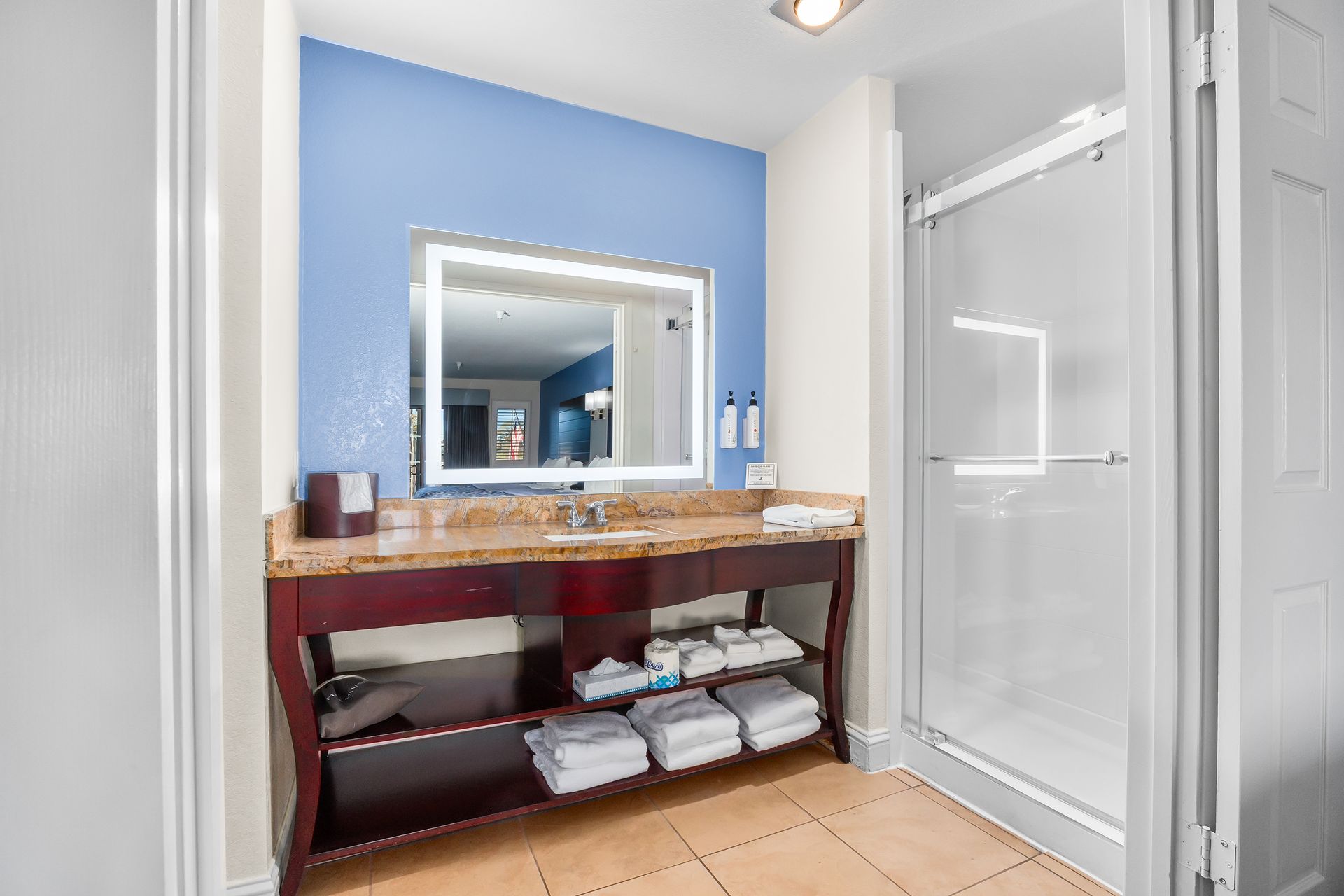 A bathroom vanity with a blue wall, a light-up mirror, granite countertop, open storage shelves, and a glass shower stall.