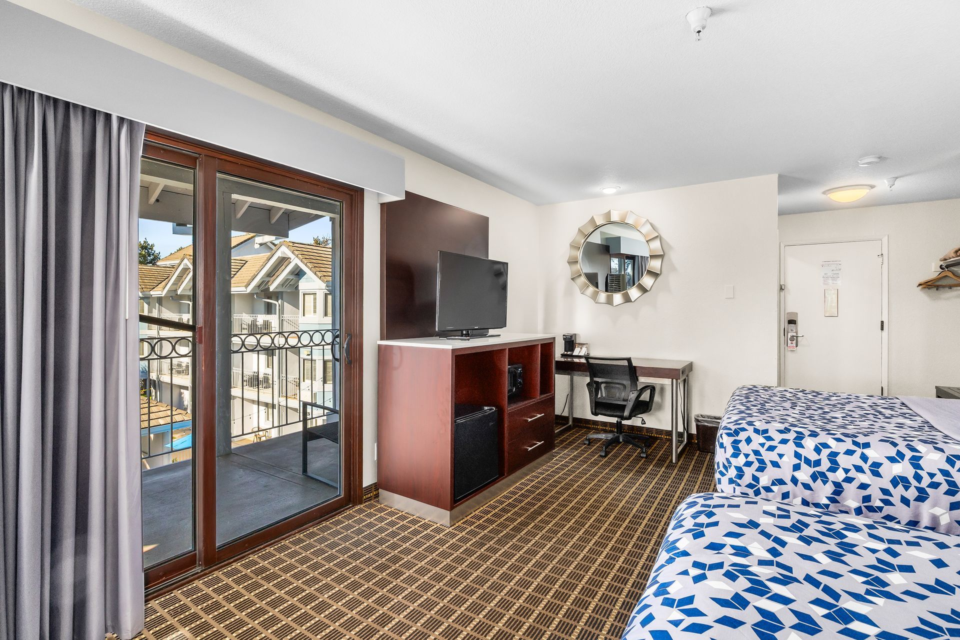A hotel room with two beds, a patterned carpet, a television on a wooden cabinet, a desk, and a balcony with a view.