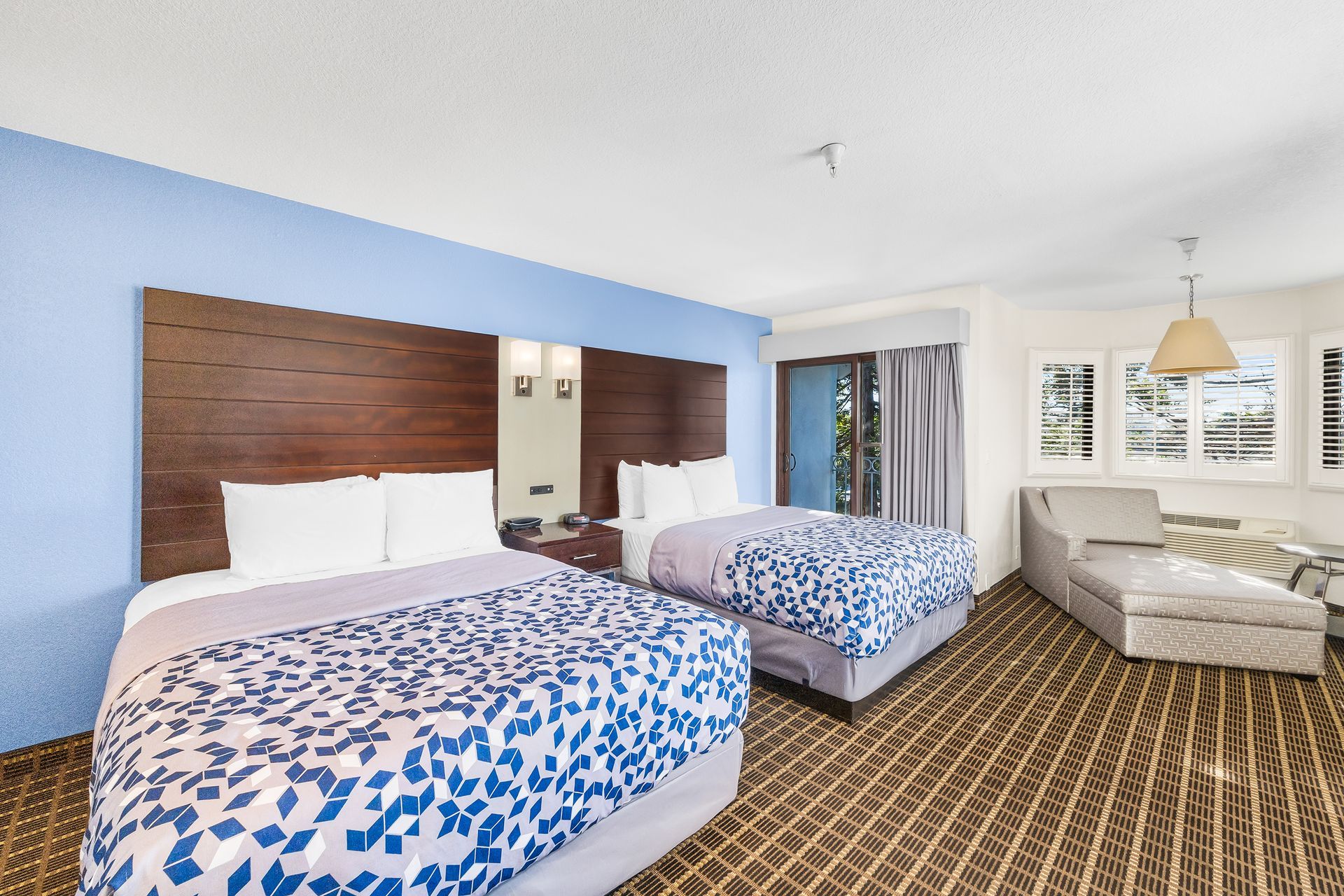 A hotel room with two queen beds, dark wood headboards, blue accent wall, and a beige chaise lounge by the window.